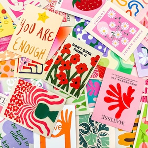 May include: A collection of colorful art prints with various designs and text. The prints feature illustrations of flowers, strawberries, and abstract shapes. Some prints have text that reads "YOU ARE ENOUGH" and "DON'T OVER THINK IT".