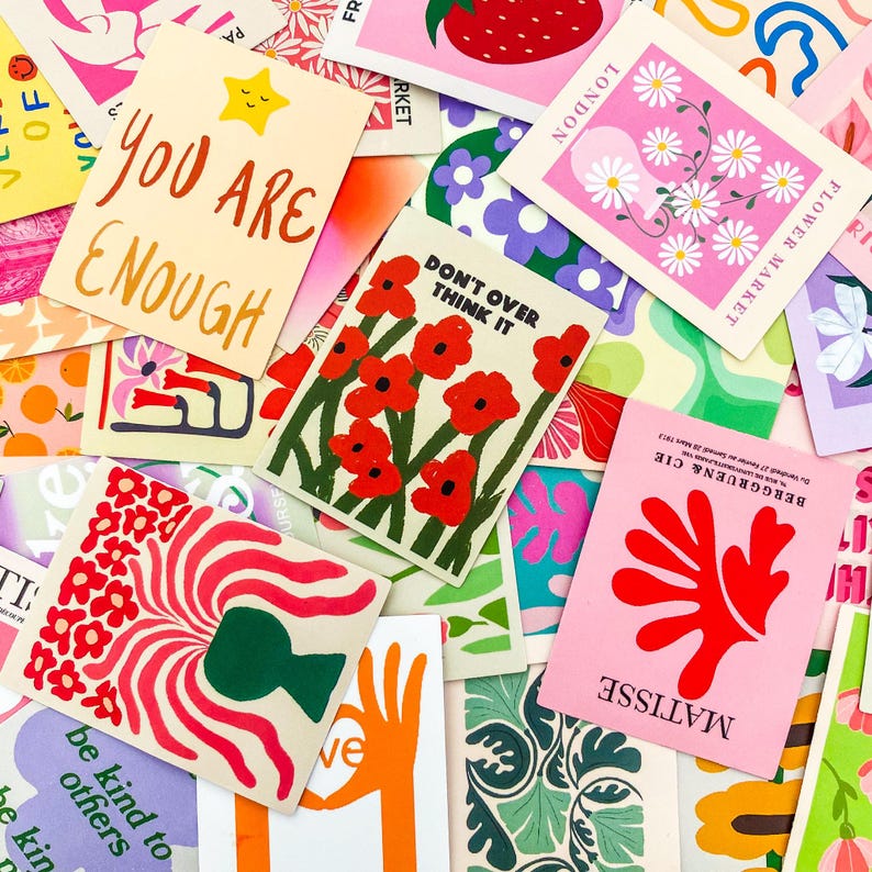 May include: A collection of colorful art prints with various designs and text. The prints feature illustrations of flowers, strawberries, and abstract shapes. Some prints have text that reads "YOU ARE ENOUGH" and "DON'T OVER THINK IT".