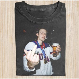 May include: Black t-shirt featuring a photo of a person holding a beer and cigarette, giving the middle finger. The person is wearing a white and blue jersey with a medal. The shirt has a vintage, worn look.