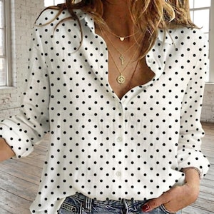 Puede incluir: Una camisa blanca con estampado de lunares negros. La camisa de manga larga tiene un cuello clásico y está parcialmente desabrochada. La modelo lleva collares dorados. La camisa es de tejido ligero.