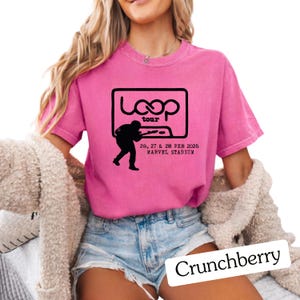 May include: A pink t-shirt with a black graphic that says "Loop Tour" with a silhouette of a person playing a sport. The text "26, 27 & 28 FEB 2025 MARVEL STADIUM" is also on the shirt. The word "Crunchberry" is at the bottom.