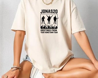 May include: A beige t-shirt featuring a black graphic with the text "JONAS20" above a band silhouette and the words "GREETINGS FROM YOUR HOMETOWN TOUR". The shirt is a casual style.