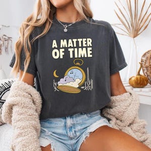 May include: Dark gray t-shirt with the text "A MATTER OF TIME" in white block letters. Below the text is a graphic of a pocket watch with a musical note, clouds, and a crescent moon.
