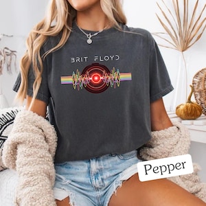 May include: Dark gray t-shirt with the text "BRIT FLOYD" above a graphic of a red and black circular design with colorful wave patterns. The word "Pepper" is written in a white box at the bottom.