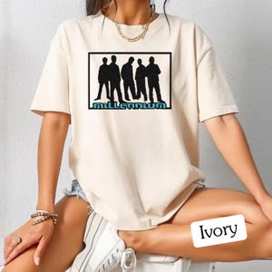 May include: Ivory-colored t-shirt featuring a black silhouette of a group of people inside a black rectangle. The word "millennium" is printed in a teal font below the silhouette. The shirt is a casual style.