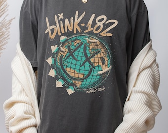 May include: Dark gray t-shirt featuring the band name "blink-182" in a distressed font above a globe design with a smiley face and the words "WORLD TOUR". The shirt has a vintage, worn look.