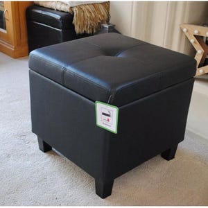 May include: A black square ottoman with a buttoned, padded top and four short legs. The ottoman is made of a dark material and has a tag attached. It is placed on a light-coloured carpet.