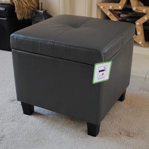 May include: A dark grey square ottoman with a buttoned top and black legs. The ottoman has a tag attached to the side. It is placed on a light-coloured carpet.