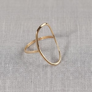 Puede incluir: Un anillo dorado con un diseño ovalado abierto único. El anillo presenta una banda fina y una forma ovalada más grande y alargada que se asienta sobre el dedo. El anillo se encuentra sobre un fondo gris neutro.