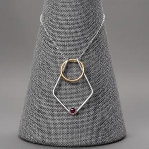 Pode incluir: Um colar de prata com um pendente geométrico. O pendente apresenta um anel dourado entrelaçado com uma forma de diamante prateado. Uma pequena pedra preciosa vermelha redonda está engastada na parte inferior do diamante.
