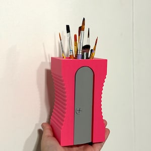 May include: A bright pink pencil sharpener-shaped desk organizer holding various paint brushes. The organizer has a gray door and a cross symbol. The brushes are of different sizes and colors, with wooden handles and metal ferrules.
