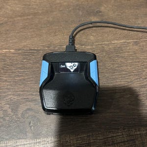 Puede incluir: Dispositivo de juego negro con detalles azules y un cable conectado. El dispositivo tiene la palabra "Zen" en la parte delantera, junto con un logotipo. El dispositivo está sobre una superficie de madera.