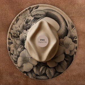 May include: A tan fedora hat with a detailed illustration of a snake and floral design. The hat features a wide brim and a rounded crown. The illustration is in shades of gray and beige, with the text "PNG Digital File" on the crown.