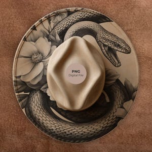 Może przedstawiać: Kapelusz fedora z beżową koroną i szerokim rondem z czarno-białą ilustracją węża i elementów kwiatowych. Kapelusz ma okrągłą naklejkę z napisem "PNG Digital File".