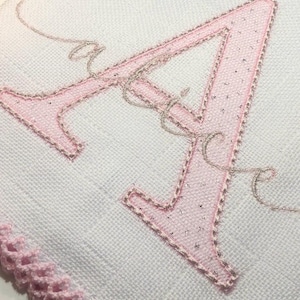 Pode incluir: Close-up de um tecido branco com uma grande letra "A" rosa bordada. A letra é contornada com fio prateado e tem uma textura brilhante. Uma delicada escrita prateada é bordada perto da letra. Uma borda rosa de crochê é visível.