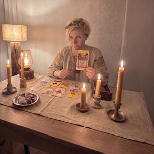 Pode incluir: Uma mulher sentada a uma mesa de madeira com cartas de tarot e velas acesas. Ela mostra uma carta de tarot com a imagem dos Amantes. A mesa &eacute; coberta com um pano bege, e uma l&acirc;mpada fornece ilumina&ccedil;&atilde;o suave.