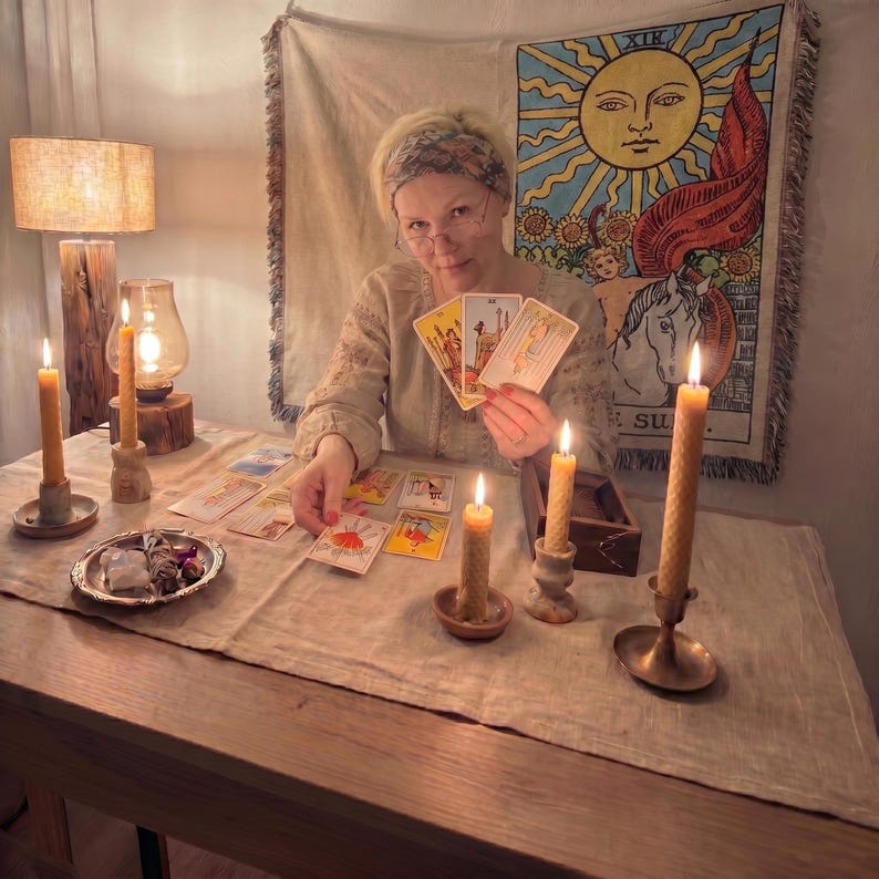 May include: A person wearing a patterned headband and beige long-sleeved shirt holds three tarot cards while sitting at a table. The table features several tarot cards, four lit beeswax candles in holders, and a small dish with crystals.