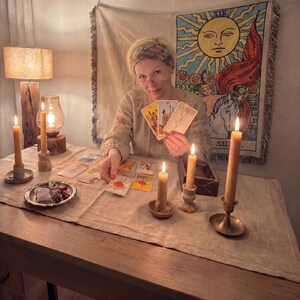 May include: A person wearing a patterned headband and beige long-sleeved shirt holds three tarot cards while sitting at a table. The table features several tarot cards, four lit beeswax candles in holders, and a small dish with crystals.