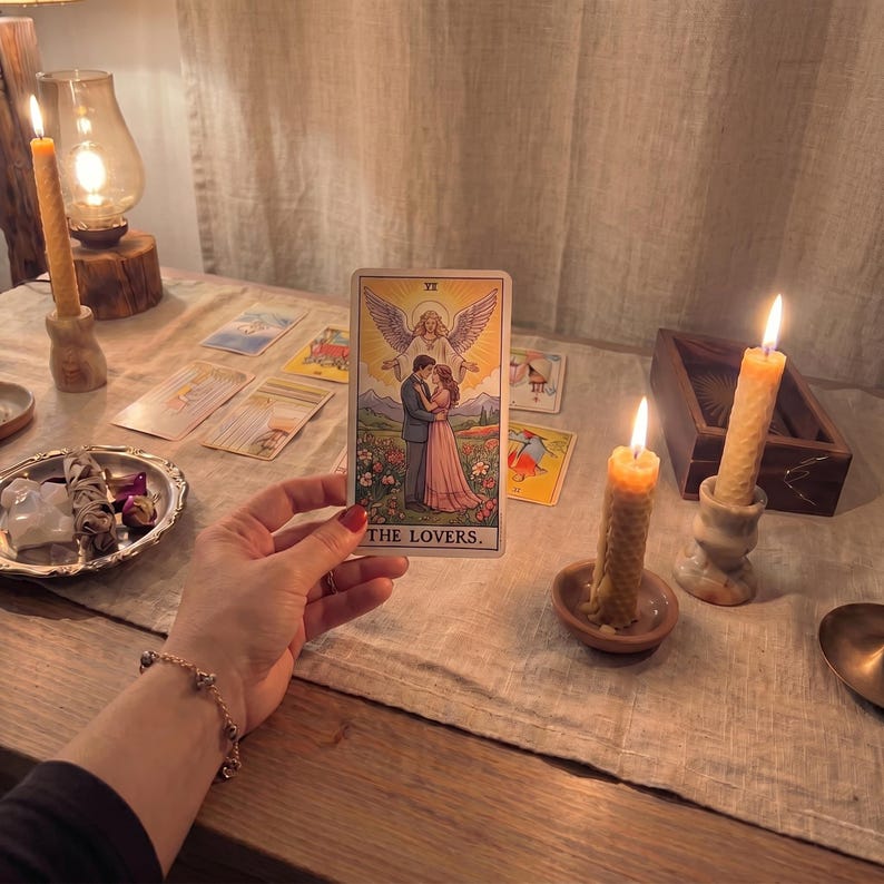 May include: A rectangular tarot card titled THE LOVERS featuring an illustration of a man and woman standing in a garden with an angel above them. The card is held upright against a background of lit beeswax candles and other scattered tarot cards.