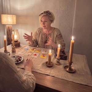 May include: A woman wearing an embroidered blouse and headscarf gestures over a table with tarot cards and lit candles. The scene is lit by a lamp and several beeswax candles, creating a mystical atmosphere.