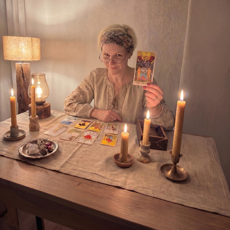May include: A tarot card reading setup featuring a deck of cards spread on a linen cloth, including The Lovers card held up. The scene includes four tall beeswax candles in various holders, a small silver dish with crystals, and a wooden box.