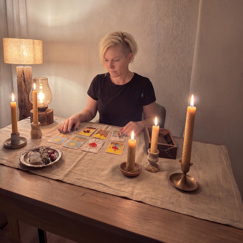 May include: A tarot card reading setup featuring a series of rectangular tarot cards laid out on a light-colored linen tablecloth. Four tall, yellow, textured beeswax candles in various holders are lit, casting a warm glow. A small silver dish holds crystals.