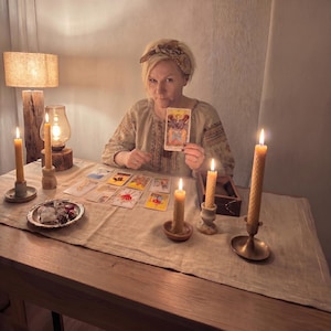 May include: A person wearing a beige patterned blouse and a patterned headband sits at a table covered with a linen cloth. Several tarot cards are spread out, and the person holds The Lovers card. Four beeswax candles and a small dish are present.