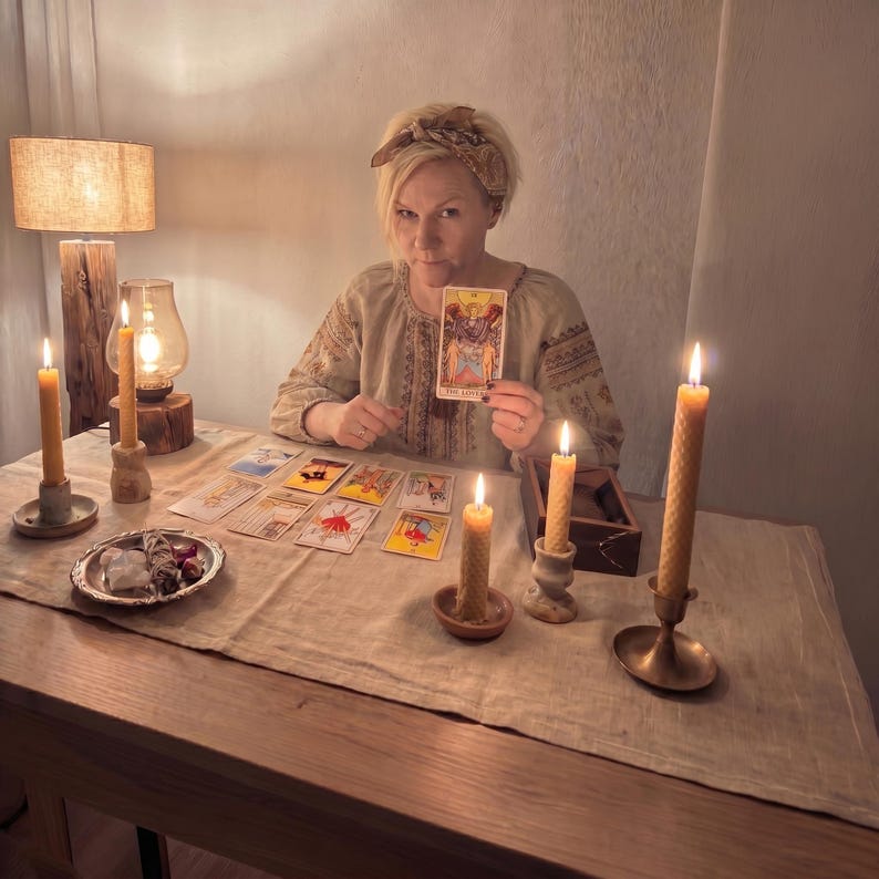May include: A person wearing a beige patterned blouse and a patterned headband sits at a table covered with a linen cloth. Several tarot cards are spread out, and the person holds The Lovers card. Four beeswax candles and a small dish are present.