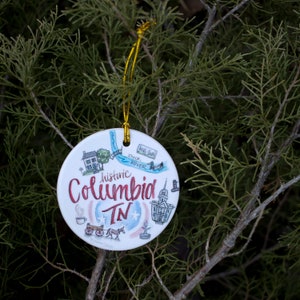 May include: A white ceramic ornament with a hand-drawn illustration of historic Columbia, TN. The ornament features a small house, a river, a bridge, a church, and a horse-drawn carriage. The text "historic Columbia TN" is written in red and black calligraphy.