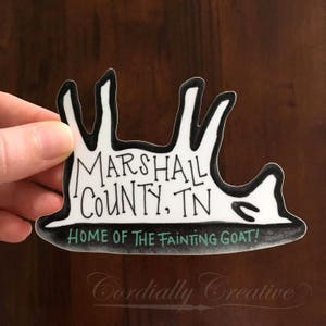 May include: Black and white sticker with a cartoon goat lying on its back. The sticker says "Marshall County, TN Home of the Fainting Goat!"