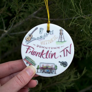 May include: White ceramic Christmas ornament with a hand-drawn illustration of downtown Franklin, Tennessee. The ornament features a guitar, music notes, a coffee cup, a water tower, a trolley, a building, and a cannon. The text "Historic Downtown Franklin, TN" is written in red and gold.