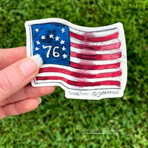 May include: A watercolor painting of a vintage American flag with 13 red and white stripes and 13 white stars in a blue canton. The number "76" is written in blue in the canton. The flag is waving in the wind.