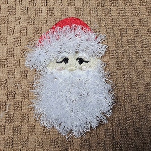 Pode incluir: Rosto do Papai Noel bordado em uma toalha marrom. O Papai Noel tem barba branca e chapéu vermelho.