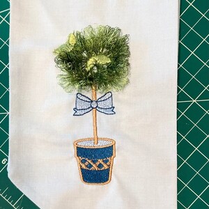 Puede incluir: Diseño bordado de un pequeño árbol verde en forma de bola con un lazo azul y una maceta marrón con detalles azules y blancos.
