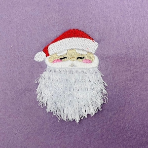 Pode incluir: Rosto de Papai Noel bordado com um chapéu vermelho e barba branca. O rosto tem um nariz rosa e os olhos fechados.