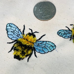 Puede incluir: Dos abejorros bordados en un fondo de tela blanco. Las abejas son amarillas y negras con alas azules. Se incluye una moneda de un cuarto de dólar estadounidense para la escala.