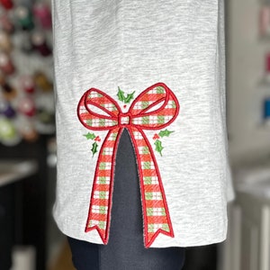 Op de afbeelding: Een grijs T-shirt met een rode en groene geruite strik die aan de zijkant is geborduurd. De strik heeft hulstbladeren en -bessen.