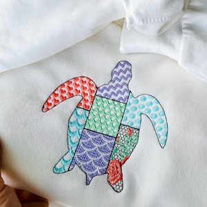 Könnte beinhalten: Ein weißer Pullover mit einem bunten gestickten Patchwork-Meeresschildkröten-Design.