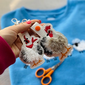 May include: Two white embroidered chicken keychains with red combs and fluffy white tails. The keychains are held in a hand against a blue background.