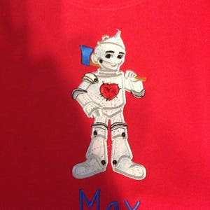 Könnte beinhalten: Rotes T-Shirt mit einem gestickten Bild des Blechmanns aus dem Zauberer von Oz. Der Blechmann hält eine Axt und hat ein Herz auf der Brust. Der Name "Max" ist unter dem Bild gestickt.