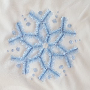 Könnte beinhalten: Ein weißer Stoff mit einem blauen gestickten Schneeflockenmuster. Die Schneeflocke ist von kleinen blauen Punkten umgeben.