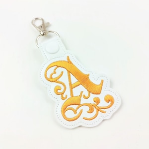 Puede incluir: Llavero de cuero blanco con una letra A bordada en oro con remolinos decorativos.