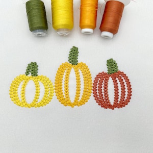 May include: Three embroidered pumpkins in yellow, gold, and orange thread are stitched onto a white fabric surface. Above the pumpkins, four spools of sewing thread in olive green, yellow, orange, and rust brown are arranged in a horizontal row.