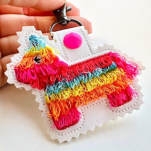 May include: A white leather keychain with a colorful, fringed pinata design. The pinata is shaped like a donkey and has a pink circle on the top.
