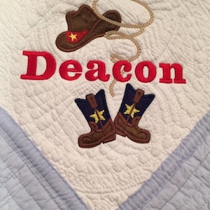 May include: A white baby quilt with a blue border. The quilt is embroidered with a cowboy hat, boots, and the name "Deacon".