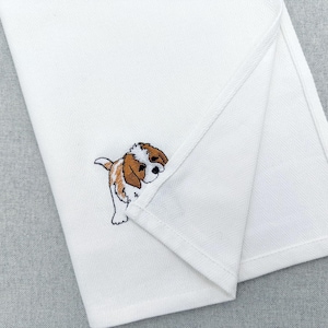 May include: White folded cloth with a detailed embroidered puppy design. The puppy is brown, white, and black. The cloth is folded to reveal the puppy peeking out from the corner.