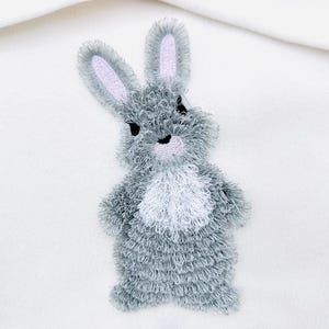 Puede incluir: Un parche bordado de un conejito gris y blanco. El conejito tiene una textura esponjosa, con pelaje gris y pecho blanco. Las orejas y la nariz están acentuadas con rosa. El conejito está de pie sobre un fondo blanco.