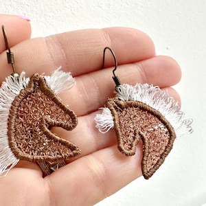 May include: A pair of horse head earrings. The earrings feature a brown, open-weave design with white fringe for the mane. The earrings are held by a person.
