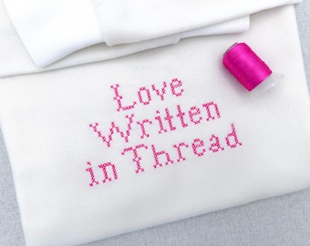 May include: A white sweatshirt featuring the text "Love Written in Thread" embroidered in a bright pink cross-stitch pattern. A small spool of matching bright pink embroidery thread is positioned on the fabric near the embroidered text.
