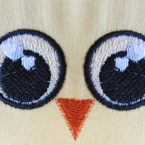 May include: Embroidered yellow fabric with two black eyes and an orange beak. The eyes are outlined in black and have white pupils.